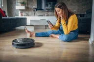 Pregnant woman using phone while robot vacuum cleaner is cleaning her home.